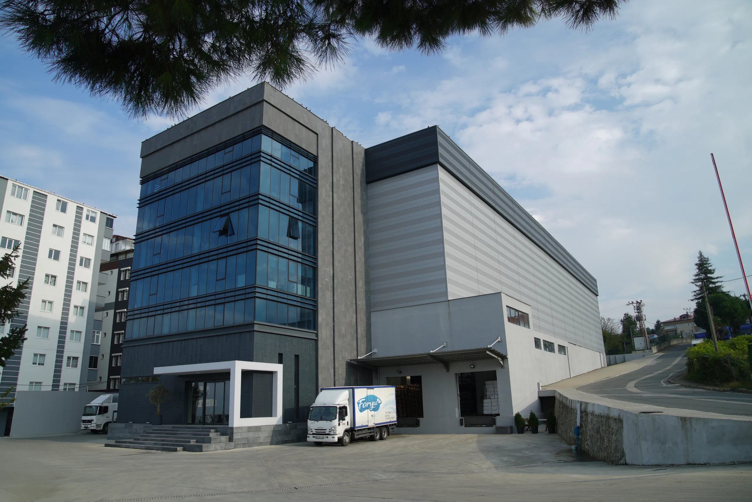 Cam cepheli koyu renkli ofis bloğu ve ona bitişik geniş gri endüstriyel hol; girişte park etmiş küçük bir dağıtım kamyonu.